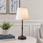 Table lamp on a round table with books and a plant in a living room setting.