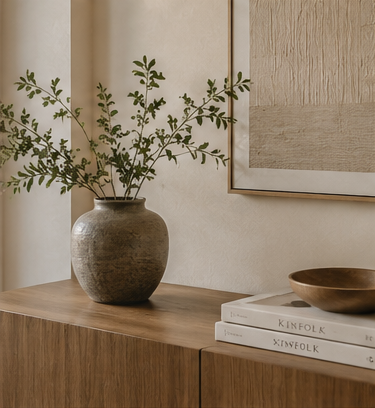 A rustic brown ceramic vase filled with leafy green branches, styled on a wooden console table next to stacked books and a wooden bowl, with a framed artwork on the wall.