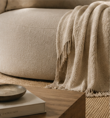 A close-up of a textured beige throw blanket draped over a round upholstered ottoman or sofa, with a wooden side table and books in the foreground on a woven rug.