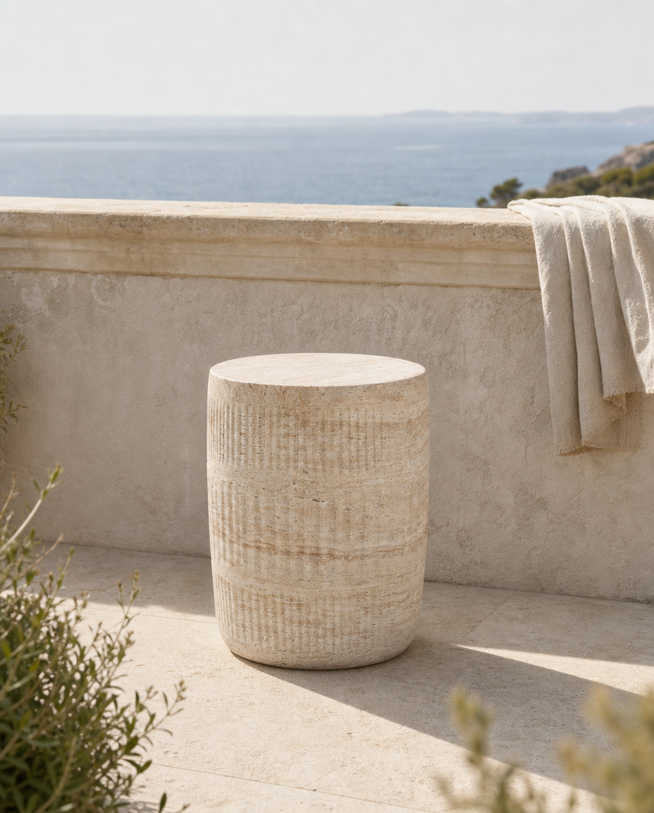 A cylindrical stone side stool with a textured, ribbed surface in a warm beige tone, set on a stone patio with a stone wall behind it and the sea in the background. A light beige towel is draped over the wall.