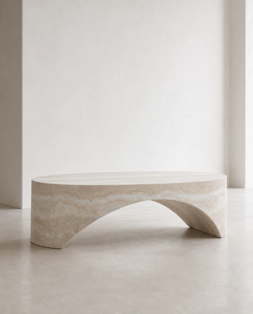 A modern curved stone bench in a light beige travertine or marble finish, set against a minimalist off-white wall and concrete floor.
