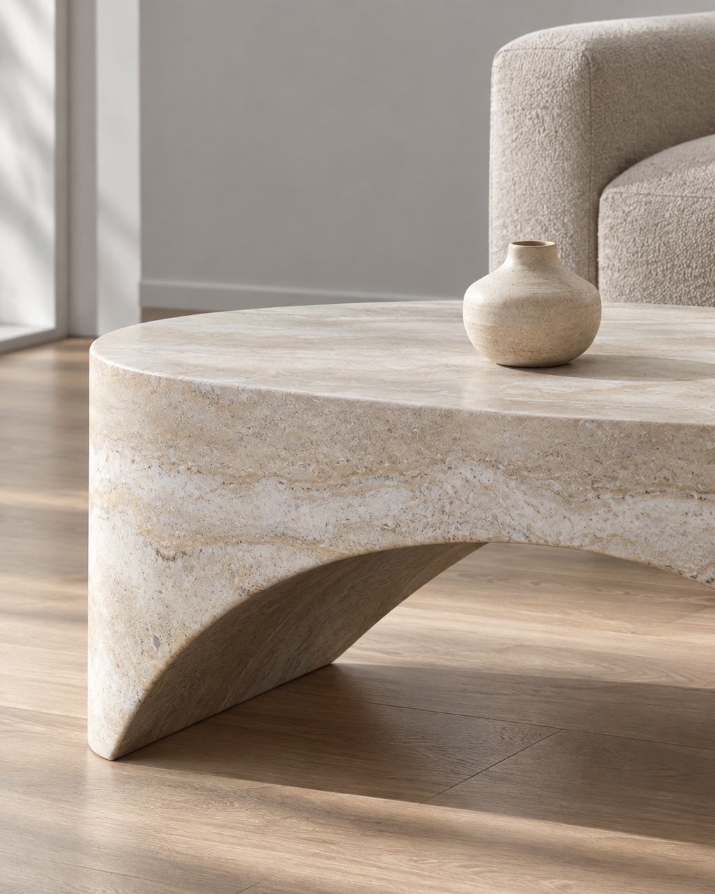 A modern round coffee table made of natural beige stone with a sculptural curved base, shown in a living room setting with a neutral sofa and a small ceramic vase on top.
