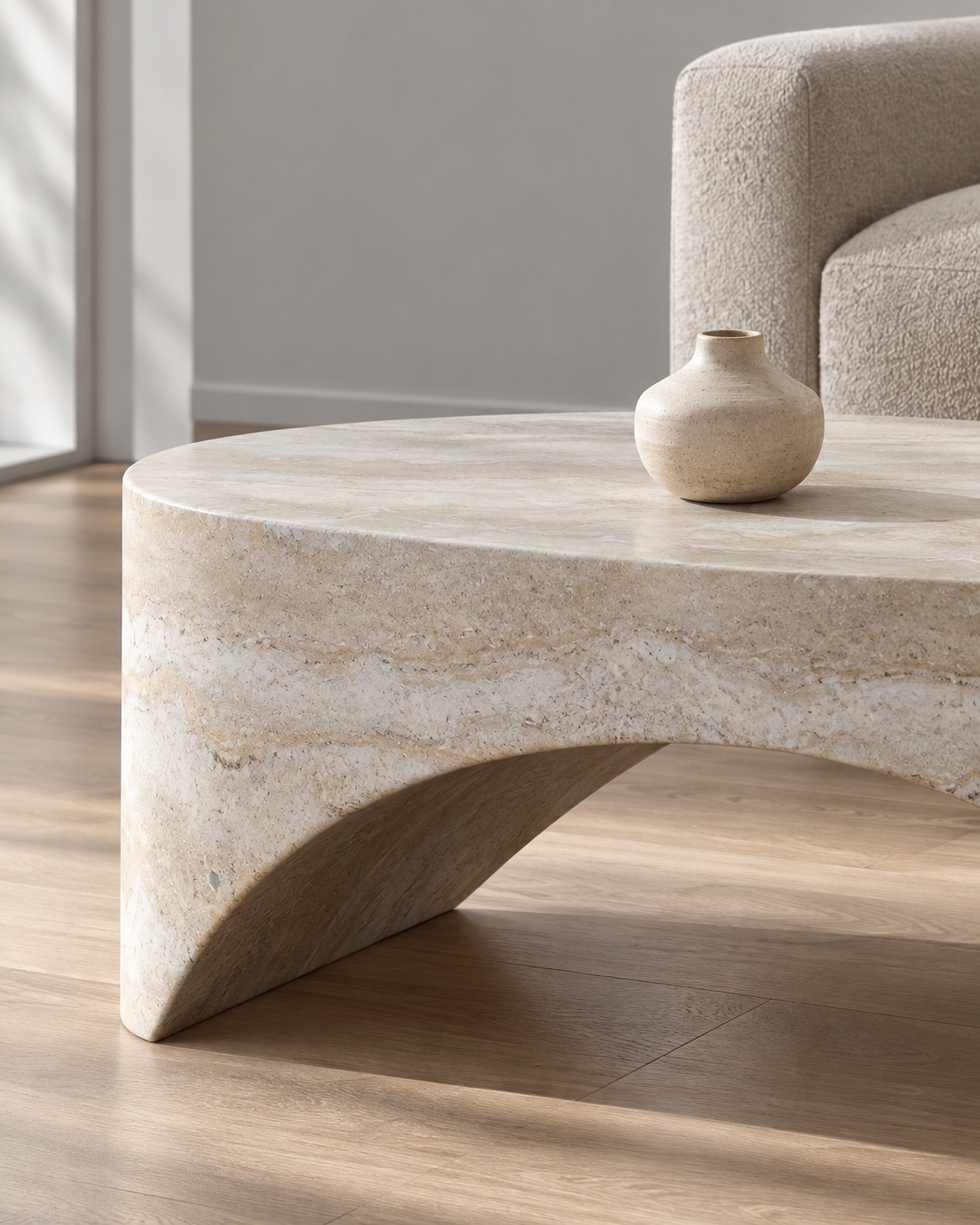 A modern round coffee table made of natural beige stone with a sculptural curved base, shown in a living room setting with a neutral sofa and a small ceramic vase on top.
