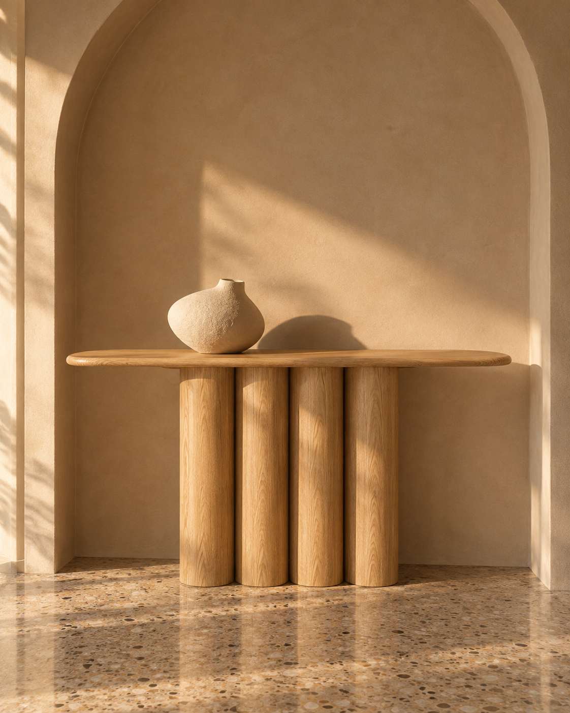A light natural wood console table with four cylindrical legs and a rounded rectangular top, styled with a small textured ceramic vase. The table is set against a warm beige plaster wall with an arched niche, receiving soft sunlight and shadows on a speckled stone floor.