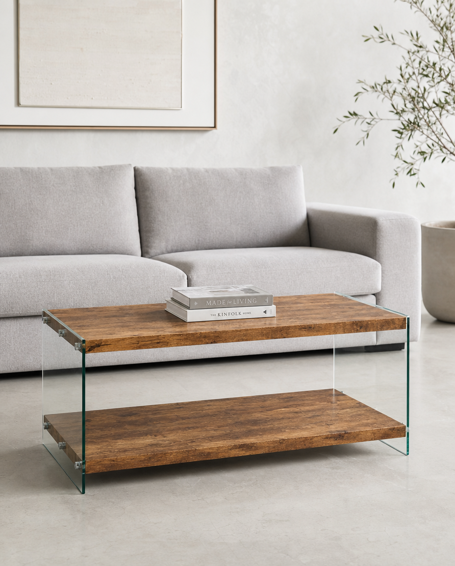 A modern two-tier coffee table with a rustic brown wood grain finish and clear glass side supports, styled in a minimalist living room setting in front of a light gray sofa.