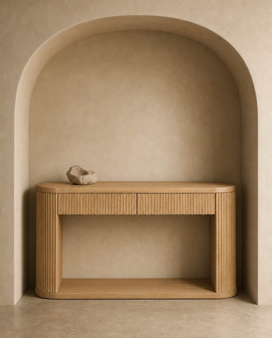 A light natural wood console table with a ribbed front and open lower shelf, placed against a warm beige plaster wall in a recessed alcove. A small stone object rests on the tabletop.