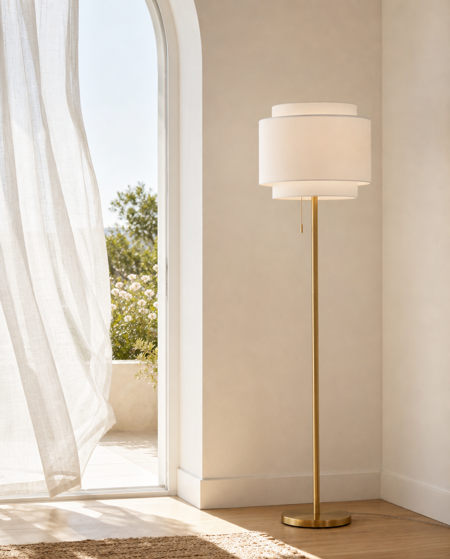 A modern floor lamp with a slim gold metal stand and two stacked white drum shades, positioned in a bright, minimalist room next to an arched window with sheer curtains.