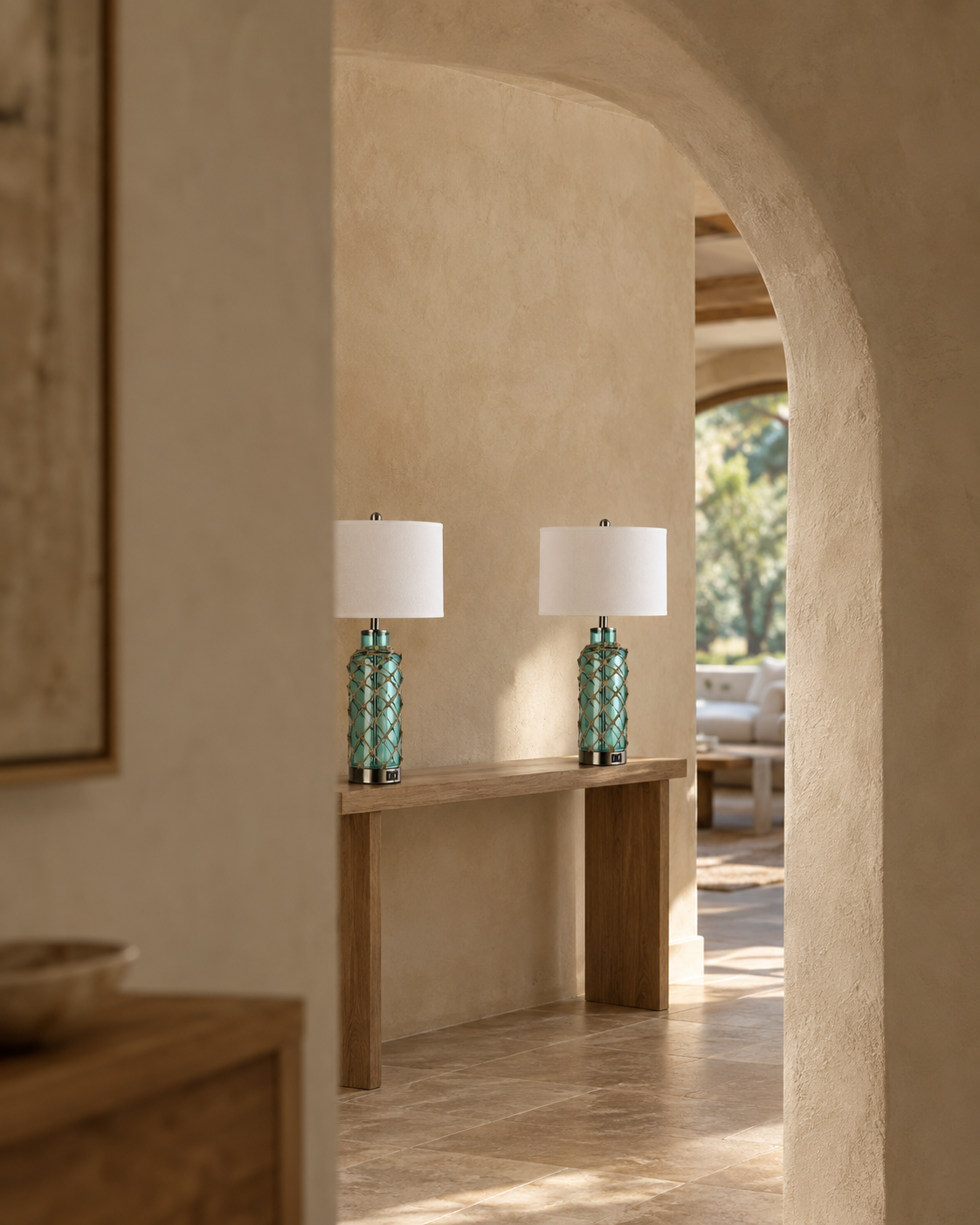 Two matching table lamps with textured turquoise glass bases and white drum shades sit on a light wood console table in a warm, arched entryway with beige plaster walls.