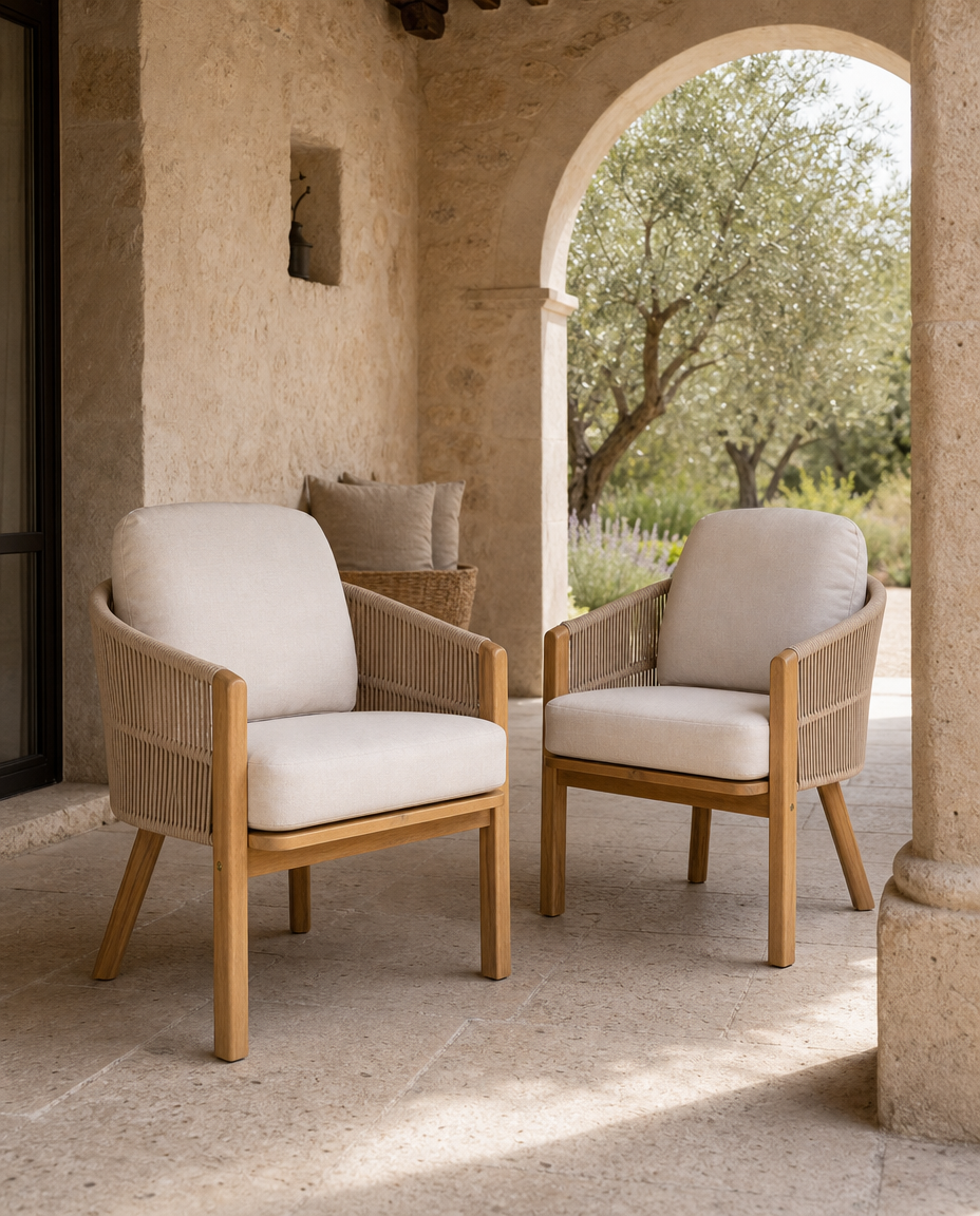 Two outdoor armchairs with light beige cushions and natural wood frames, positioned on a stone patio under a rustic archway with an olive tree in the background.