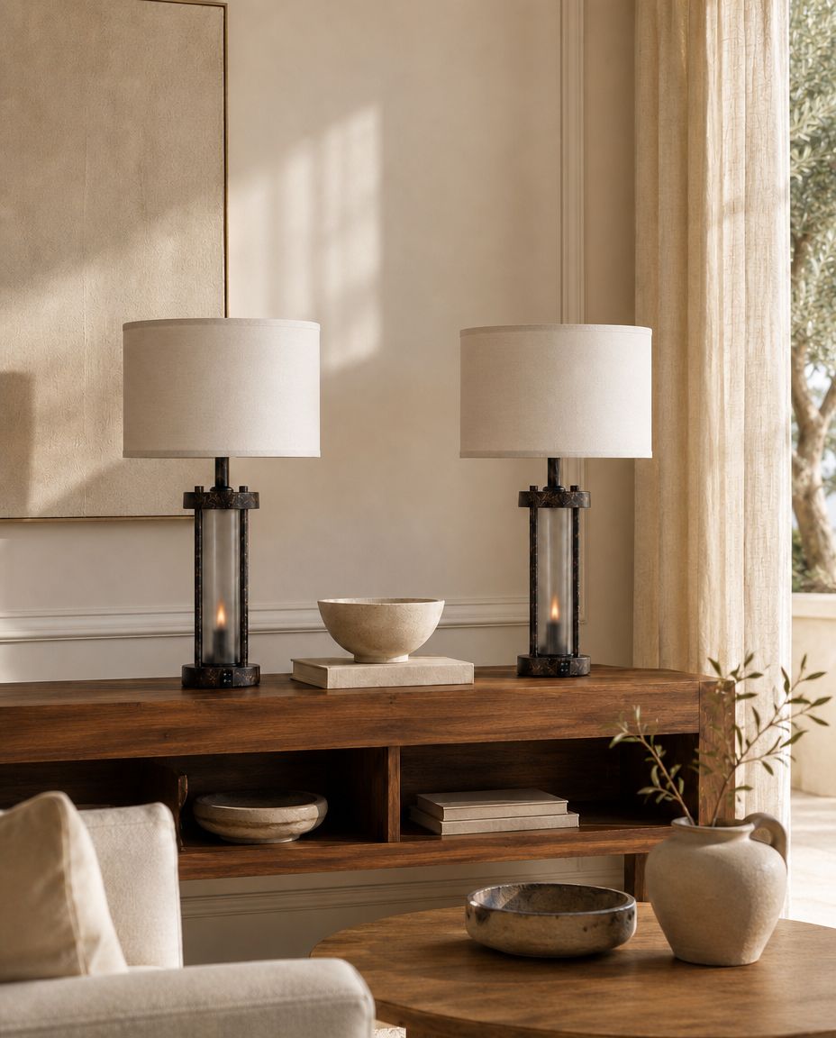 Two matching table lamps with white drum shades and black metal bases sit on a wooden console table in a softly lit living room. The scene includes neutral decor, a chair, a round table, and curtains by a window.
