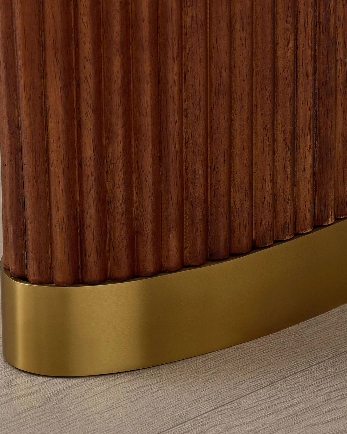 Close-up of a modern furniture piece with a fluted dark brown wood surface and a curved brushed gold metal base on a light wood floor.