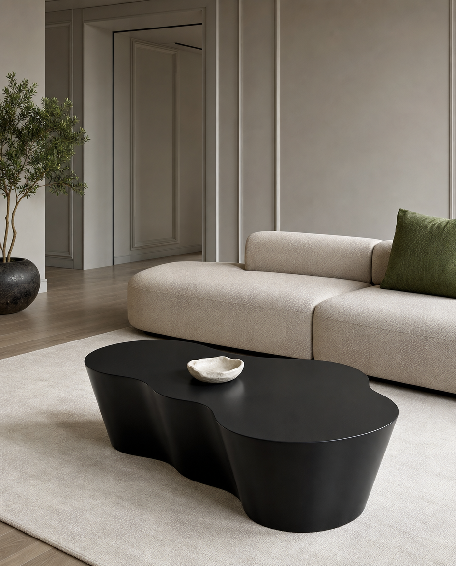 A modern black organic-shaped coffee table in a minimalist living room setting with a beige sofa, a green accent pillow, and a light beige rug.