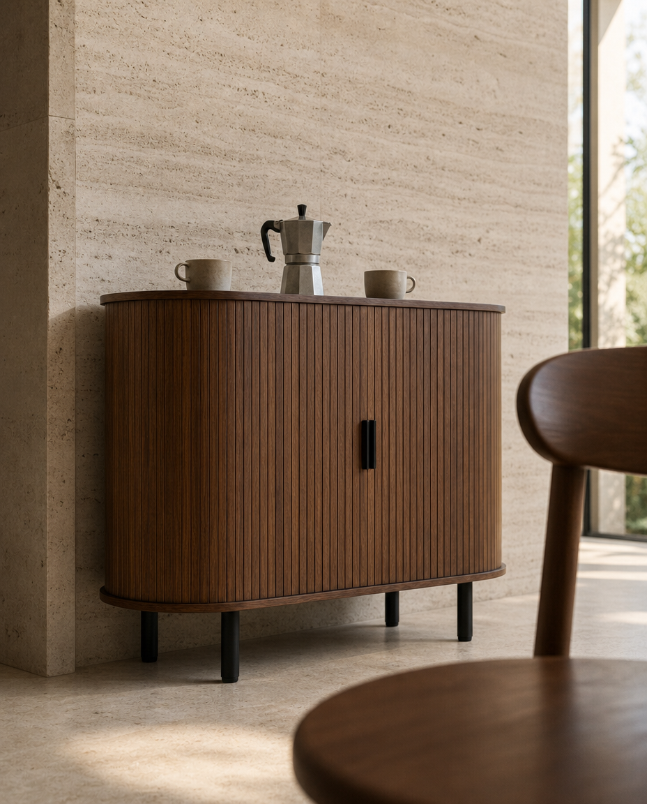 A mid-century modern wooden credenza or sideboard cabinet with vertical fluting, black legs, and a dark wood top, styled in a minimalist interior with a coffee maker and cups on top.