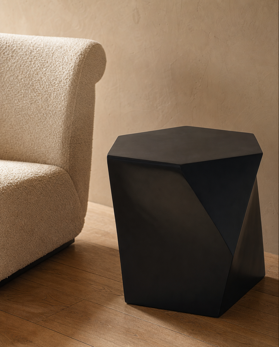 A modern black geometric side table with a faceted, diamond-like design, placed on a wooden floor next to a textured cream boucle armchair against a warm beige wall.