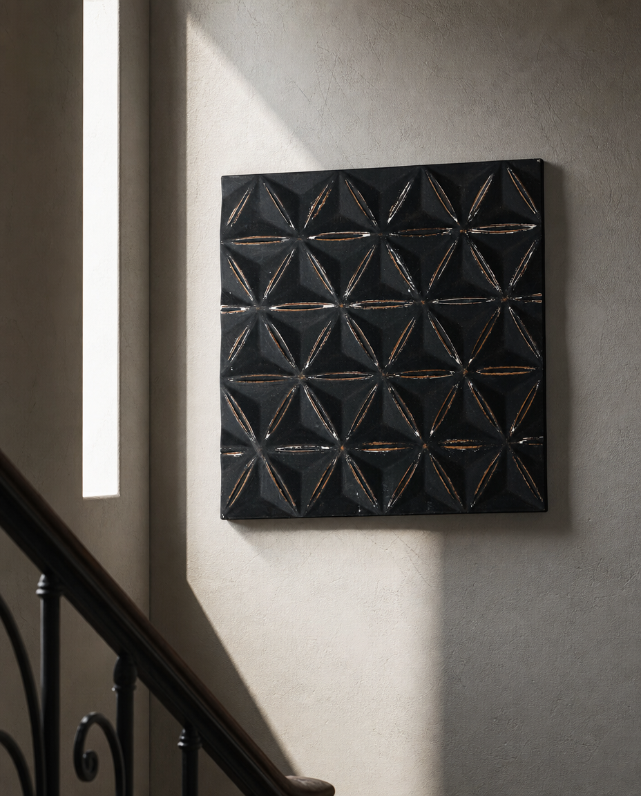 A square black textured wall art piece with a raised geometric star pattern, hanging on a textured beige wall next to a staircase with a dark metal railing.