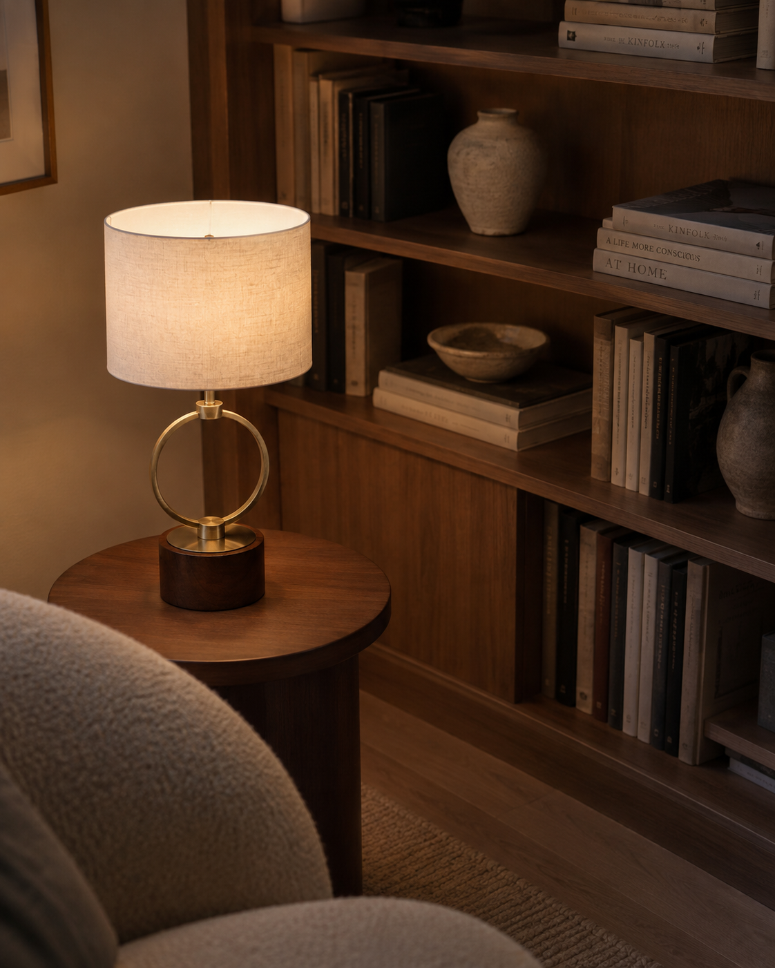 A lit table lamp with a white drum shade and gold circular metal base sits on a round wooden side table in a cozy room with a bookshelf and upholstered chair visible.